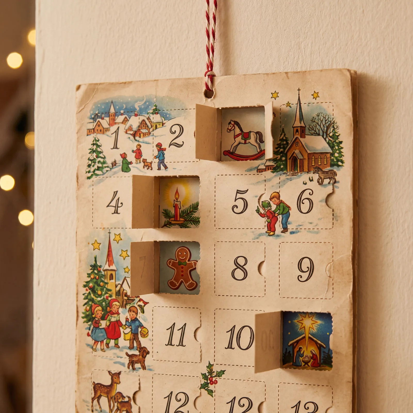 Close-up of a vintage 1960s paper Advent calendar with small numbered doors, some open, hanging on a wall, warm indoor l