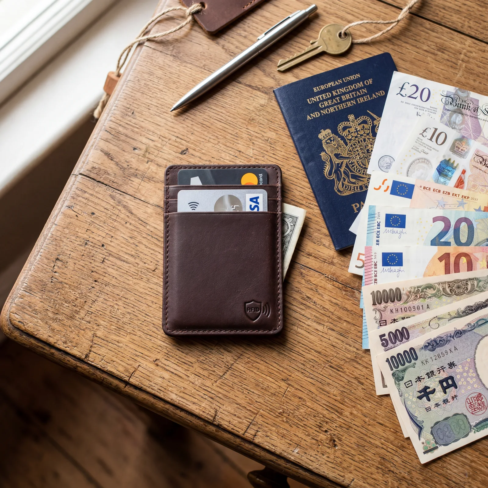 A slim card wallet with an RFID-blocking shield symbol next to a stack of foreign currency and a passport, flat lay on a wooden surface, photorealistic, warm editorial photography, no text, no watermark, 16:9