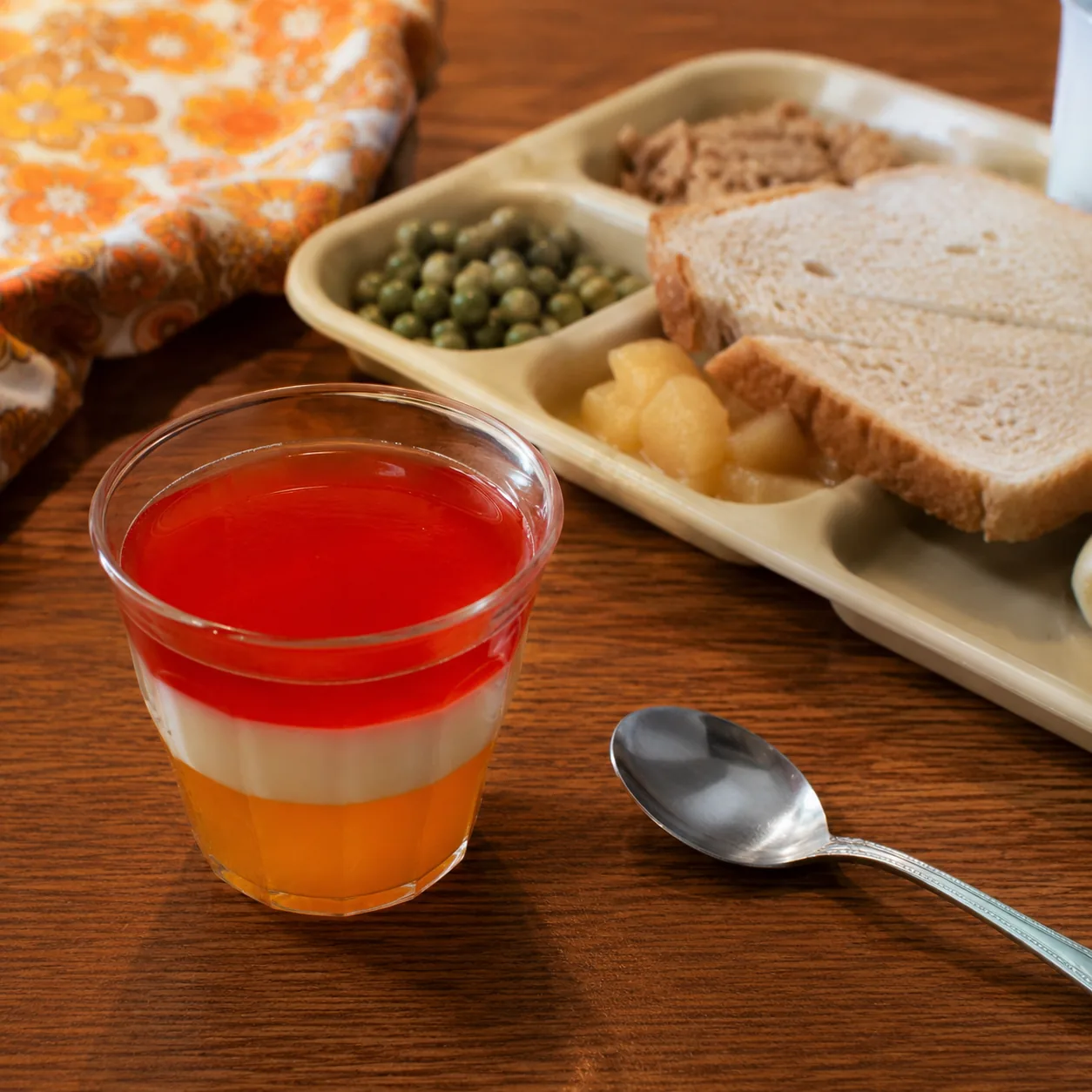 Realistic editorial photo of a layered gelatin dessert in a small glass cup beside a school lunch tray and spoon, practi