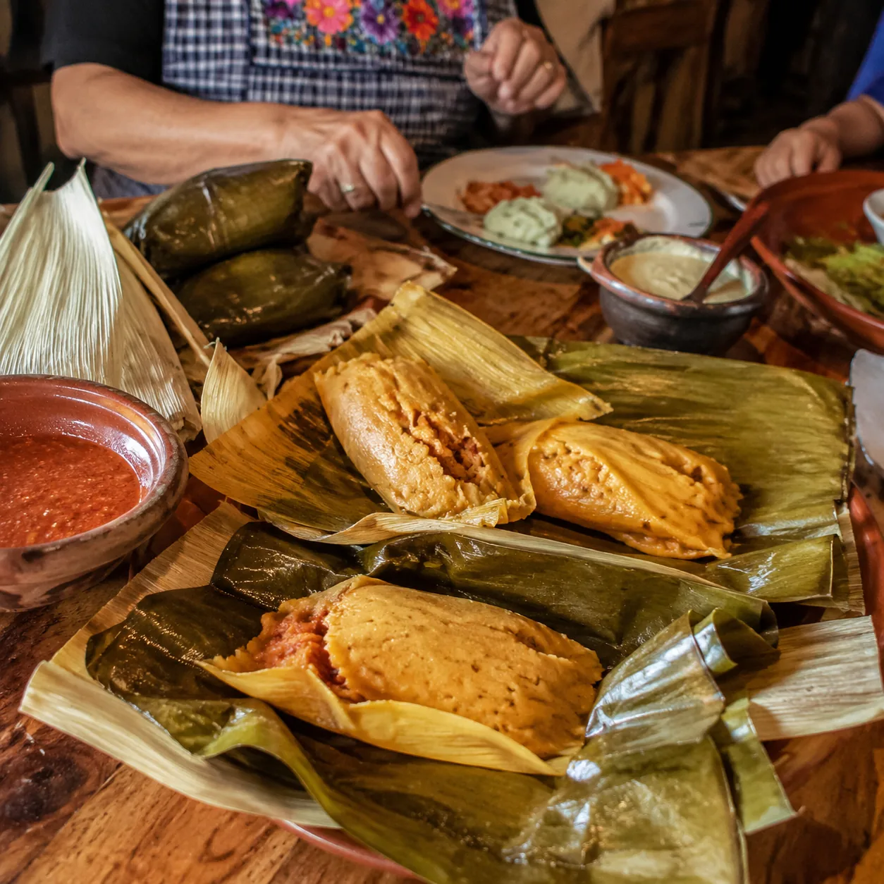 Realistic editorial photo of unwrapped tamales on banana leaves and corn husks beside salsa and family plates, warm natu