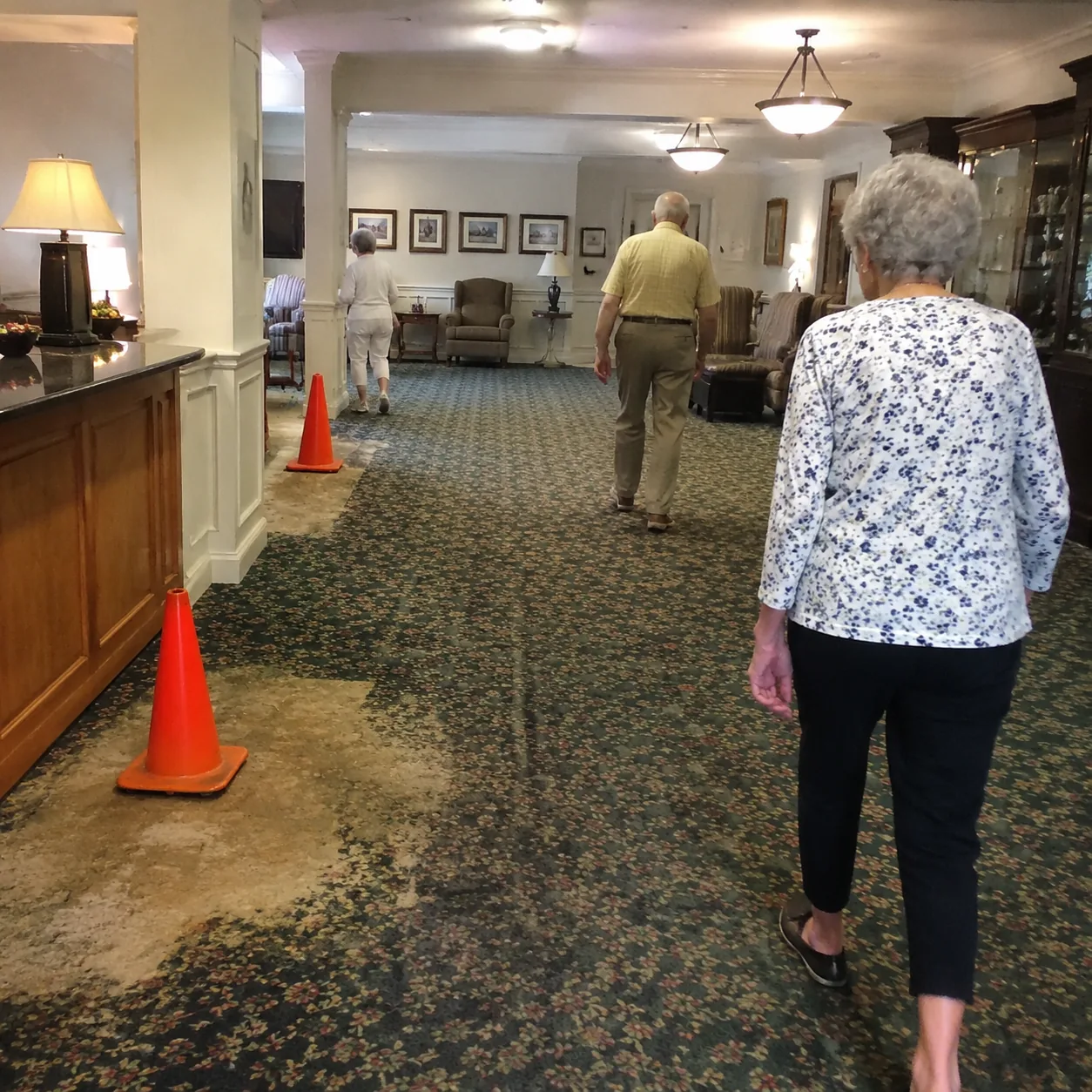Realistic editorial photo of a dated retirement community clubhouse lobby with older residents walking past worn carpet