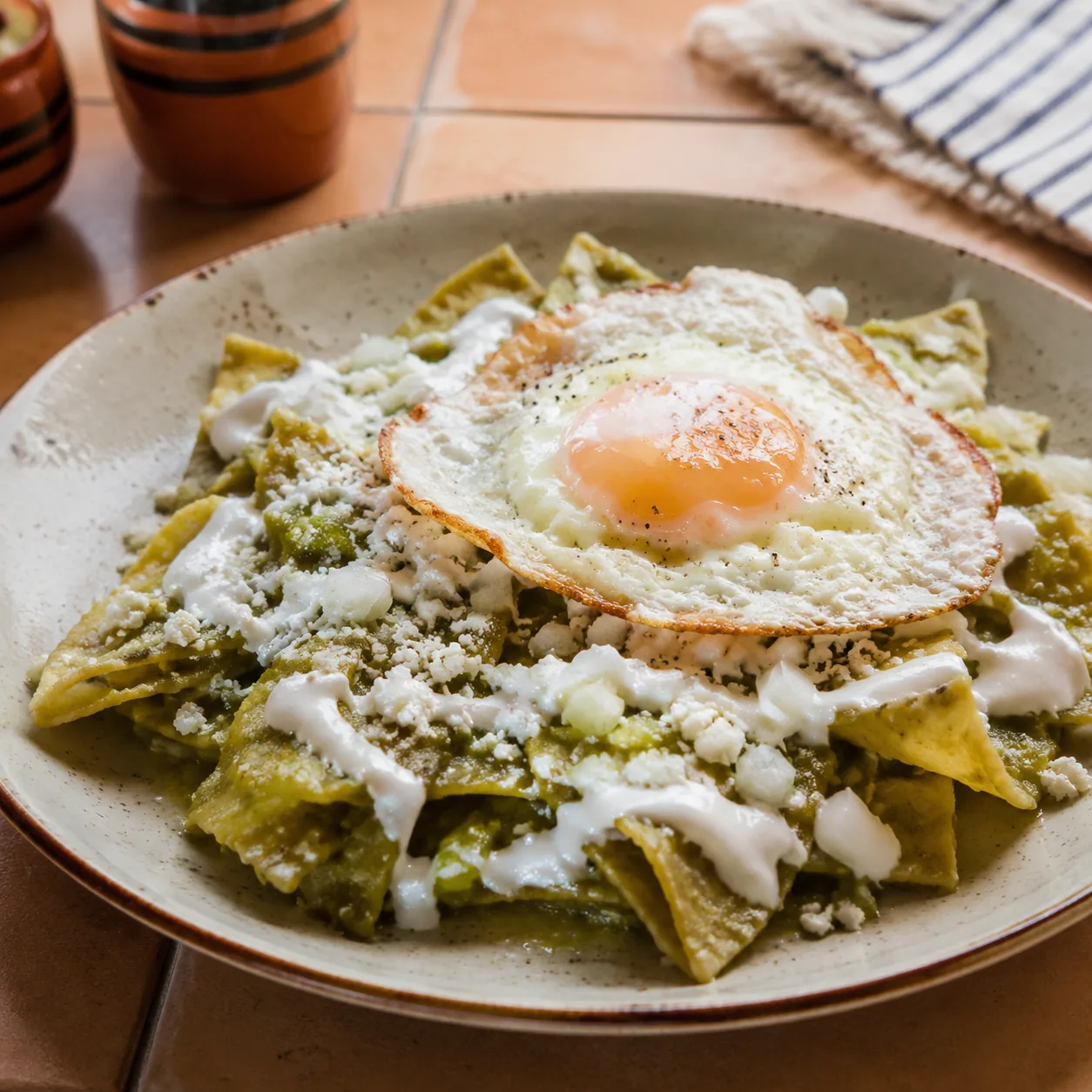 Realistic editorial photo of chilaquiles with salsa verde, crema, queso, onion, and fried egg on a breakfast plate, warm