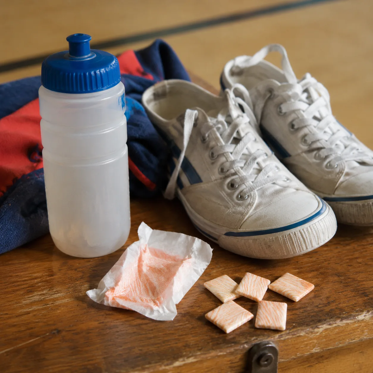 Realistic editorial photo of flat citrus chewing gum pieces beside a sports bottle and school gym shoes, practical 1980s