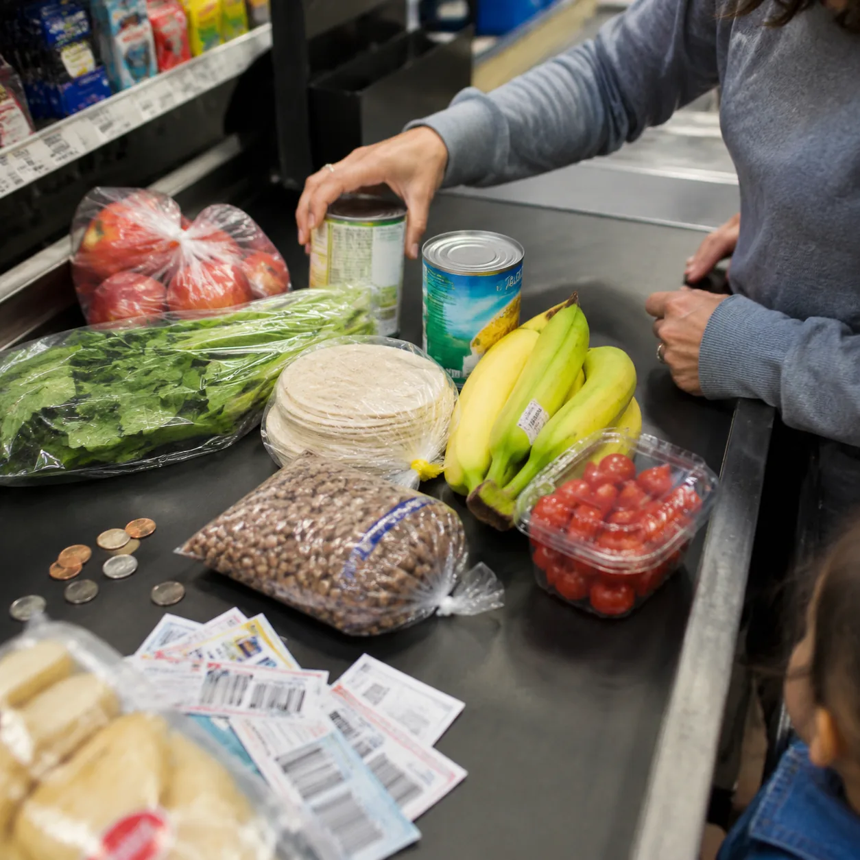 Realistic editorial photo of a grocery checkout belt with coupons, coins, produce, tortillas, and pantry staples being a