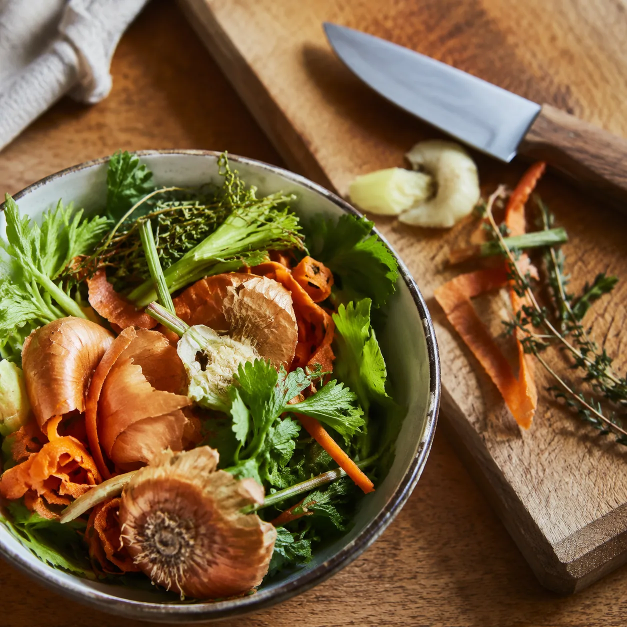 Realistic editorial photo of carrot peels, celery leaves, onion ends, and herb stems collected in a bowl beside a cuttin