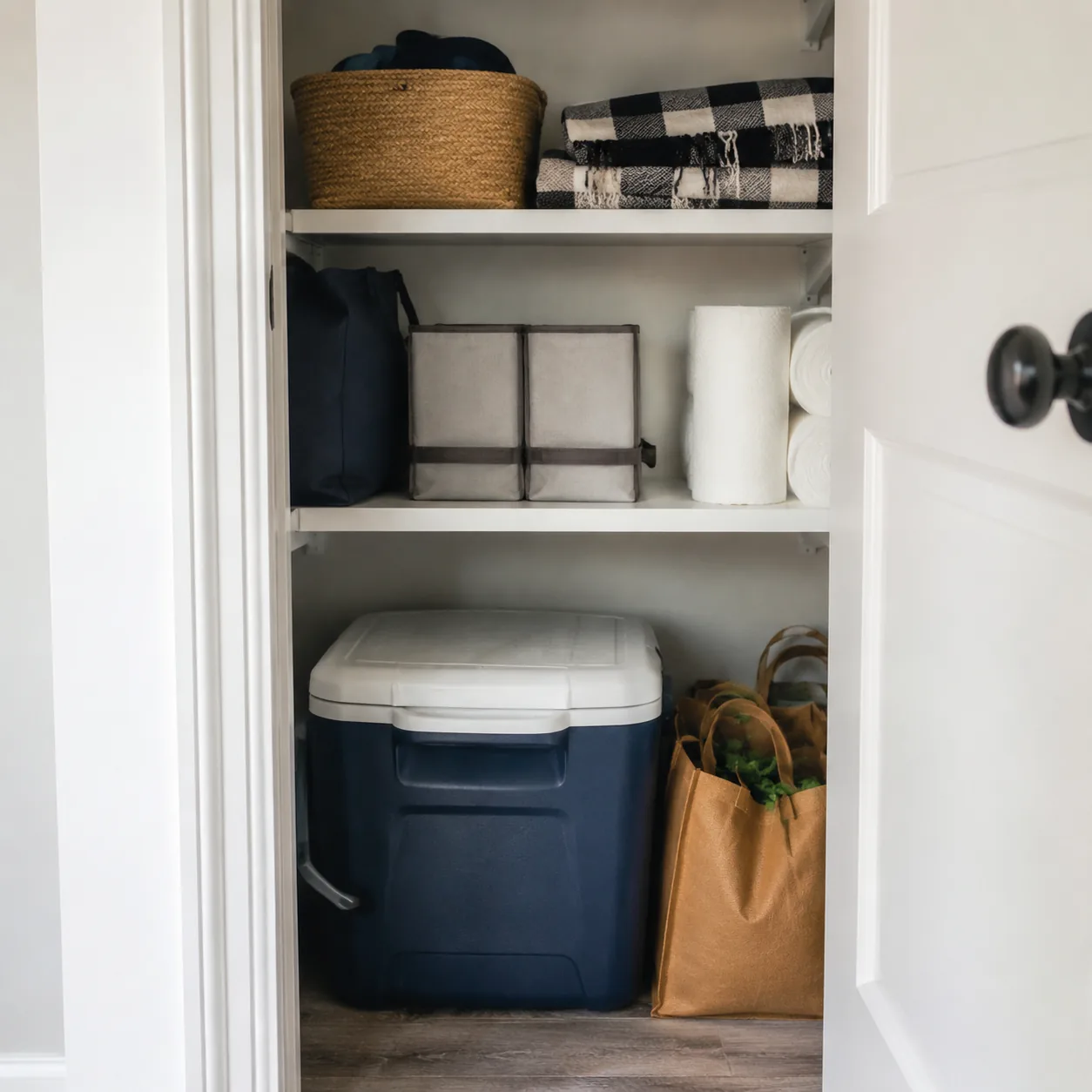 Realistic editorial photo of a medium cooler stored neatly in a utility closet with picnic blanket and grocery bags, pra