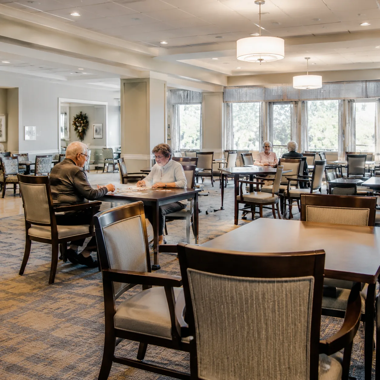 Realistic editorial photo of a retirement community activity room with a few residents at tables and many empty chairs,