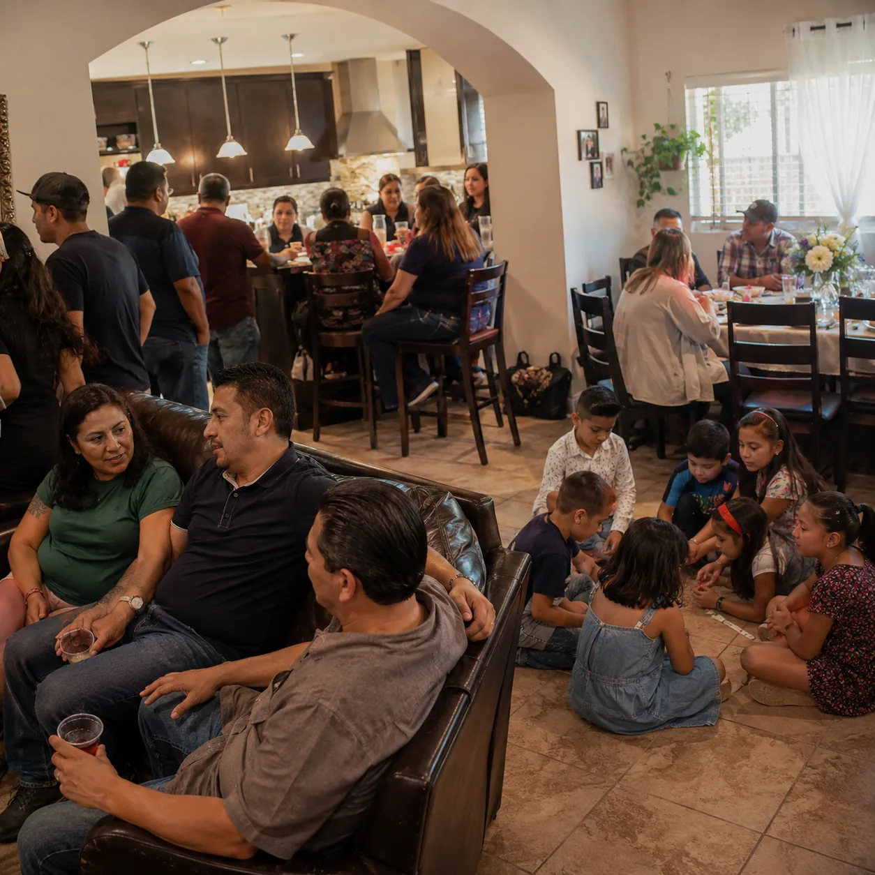 Realistic editorial photo of children and adults gathered in separate corners of a busy Latino family party at home, war