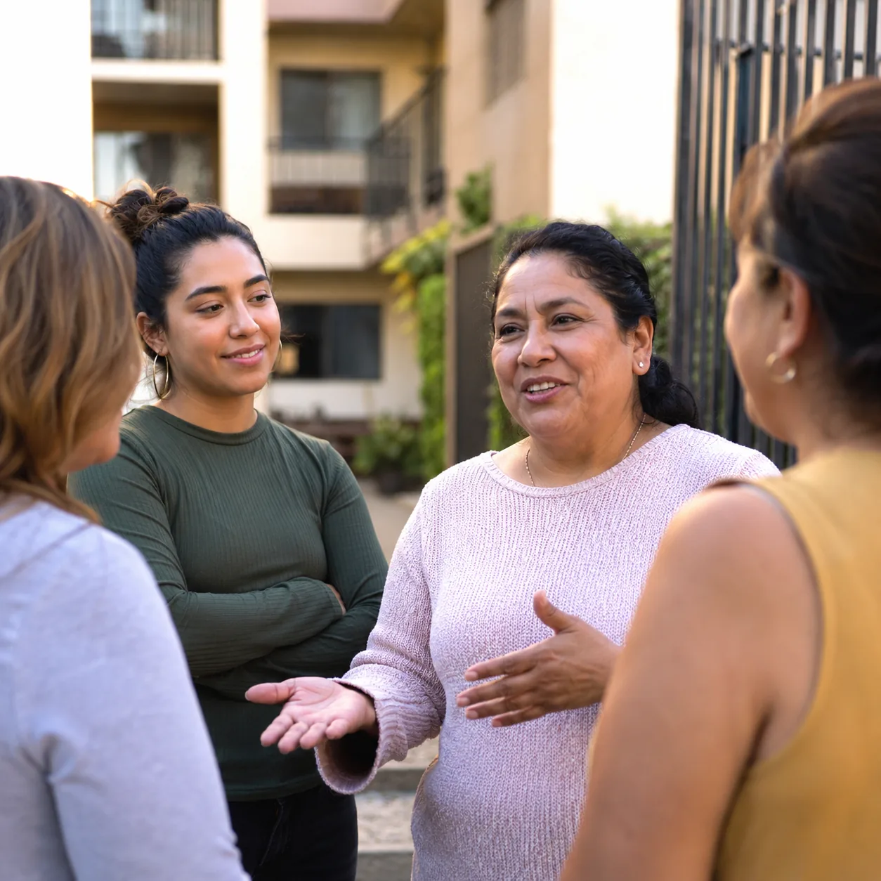 Realistic editorial photo of a Latina mother chatting with neighbors outside an apartment building while an adult child