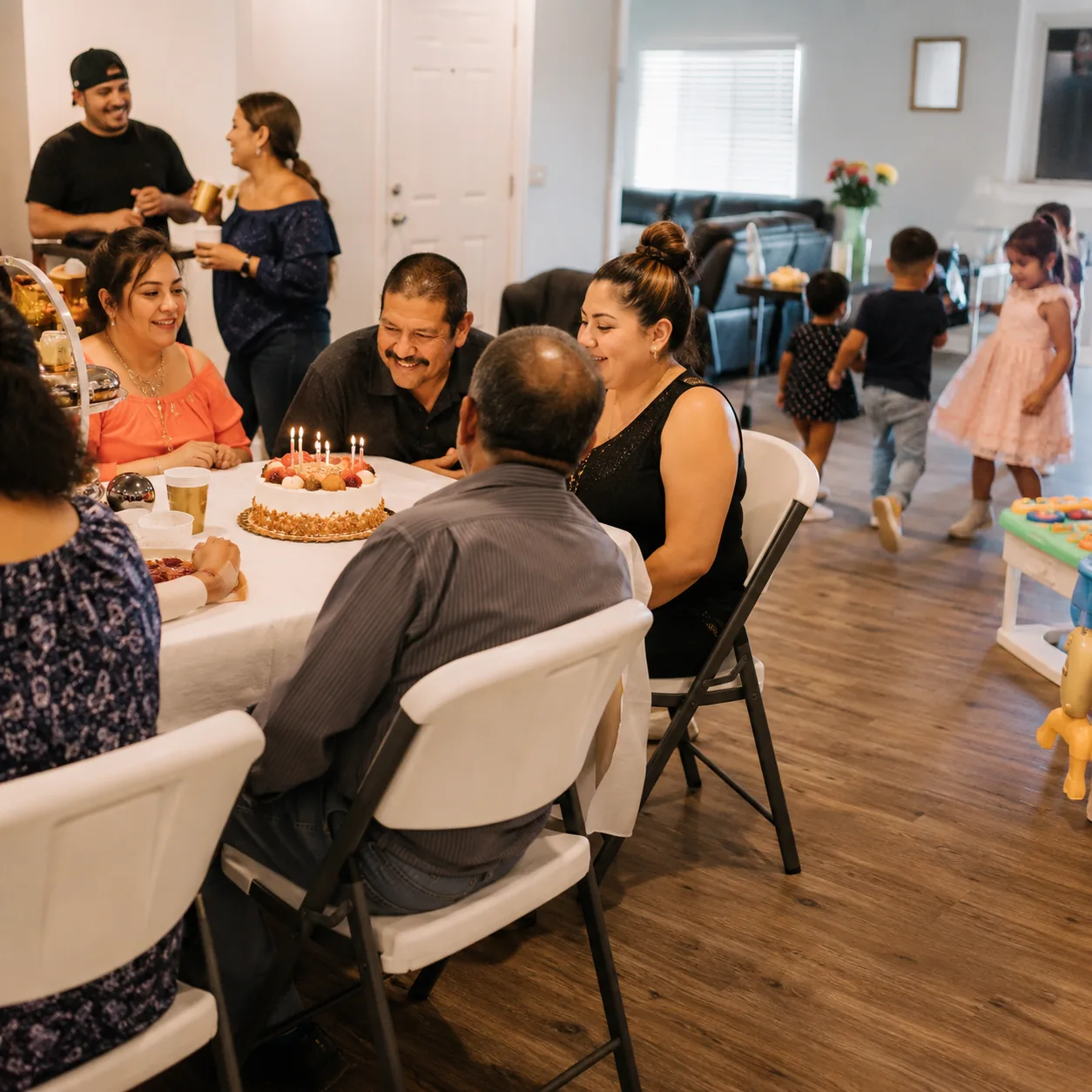 Realistic editorial photo of a Latino family birthday party at home with cake, relatives, folding chairs, and kids playi