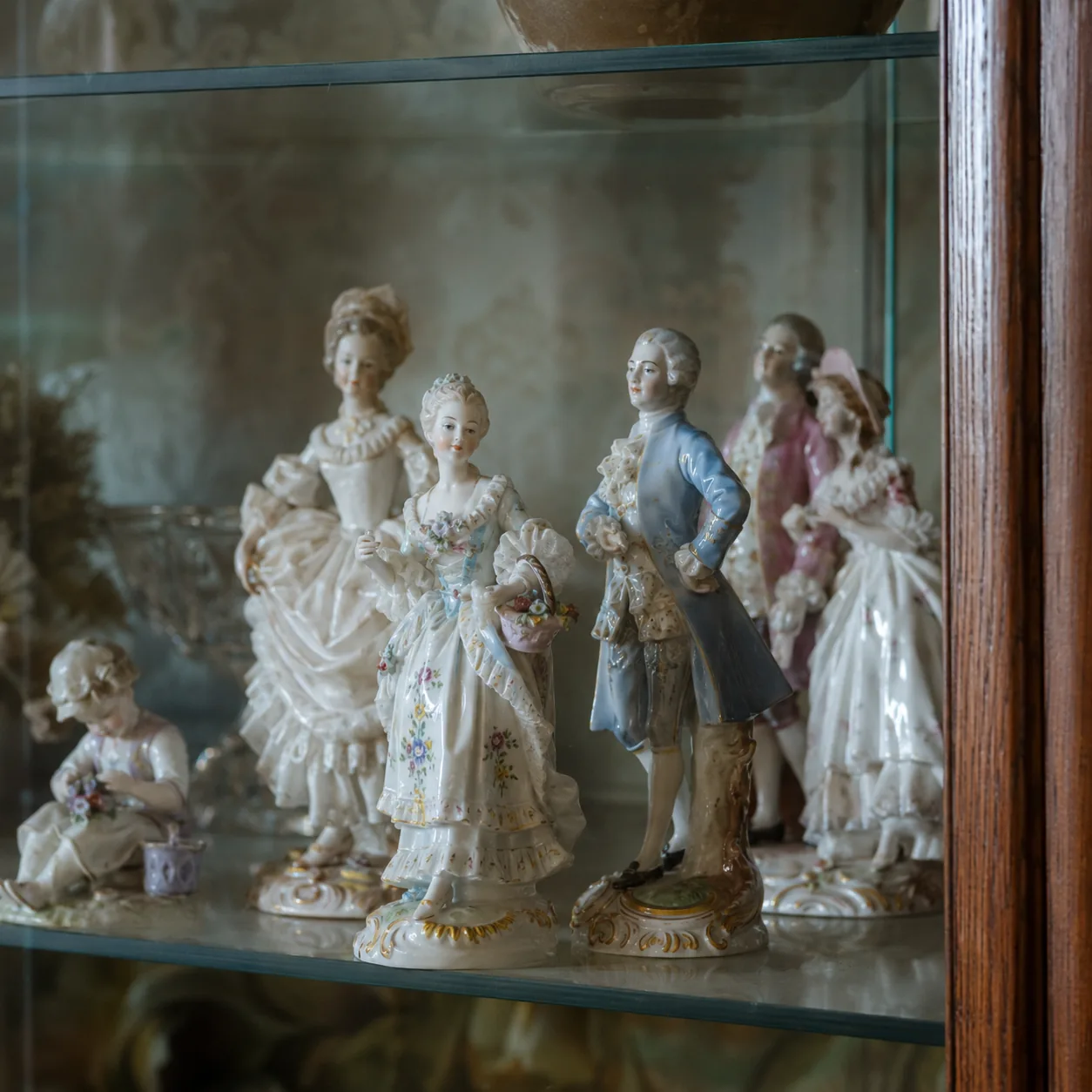 Realistic editorial photo of delicate porcelain figurines displayed on a glass shelf in an old living room cabinet, soft