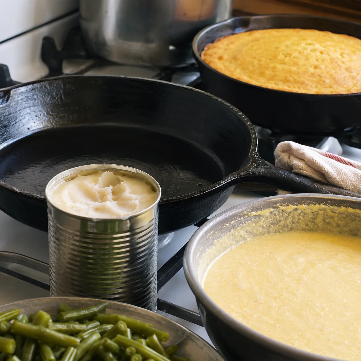 Realistic editorial photo of a clean metal can of bacon grease beside a cast iron skillet, cornbread batter, and green b
